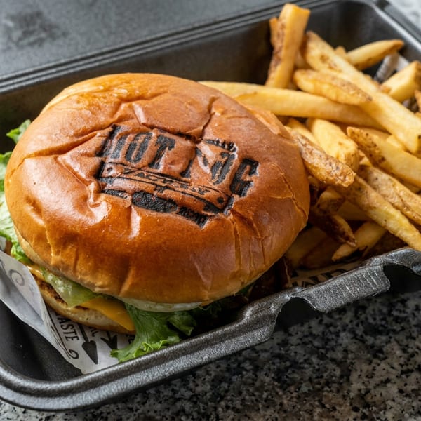 Grilled burger with lettuce and cheese beside golden fries in a black takeout tray on a granite countertop.
