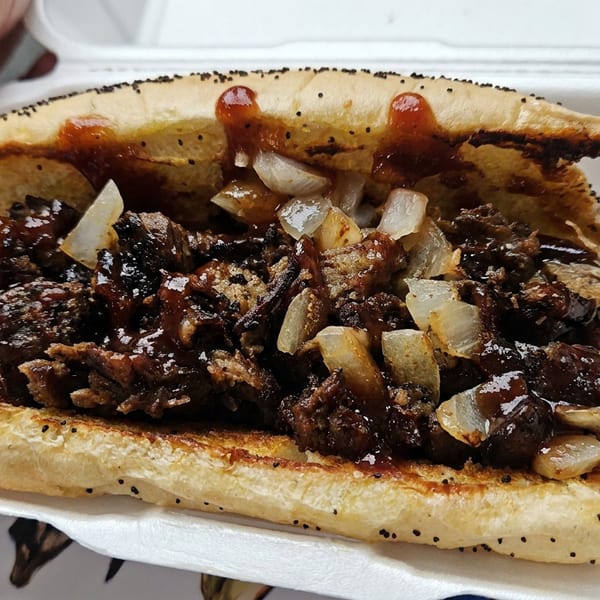 Grilled meat sandwich with chopped onions and brown sauce served in a foam takeout container.