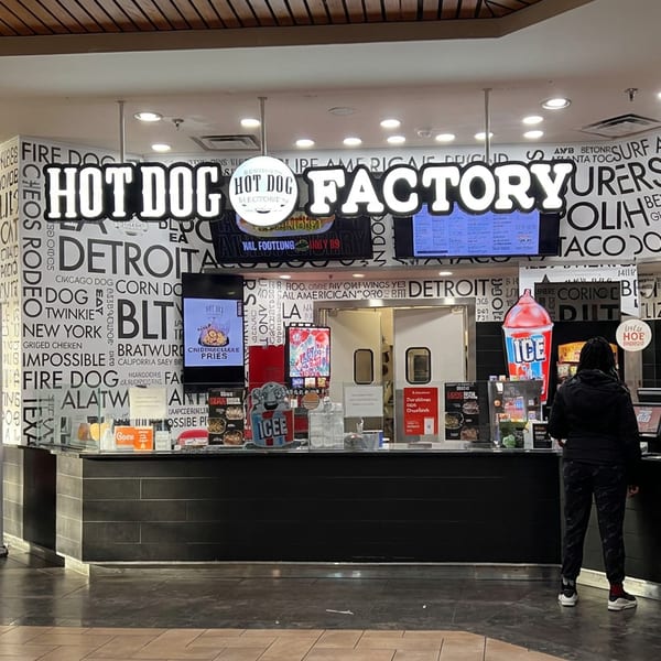Hot Dog Factory restaurant counter featuring colorful menu boards, hot dog branding, frozen drinks, and a customer ordering.