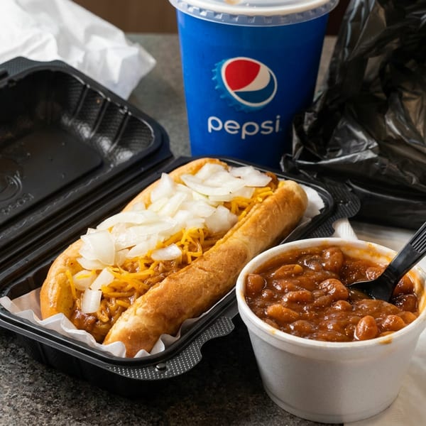 Takeout hot dog topped with shredded cheese and onions, served with baked beans, Pepsi, napkins, and packaging.