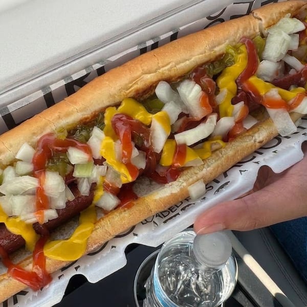 Loaded chili dog in a toasted bun topped with mustard, ketchup, relish, and diced onions, held over a tray.