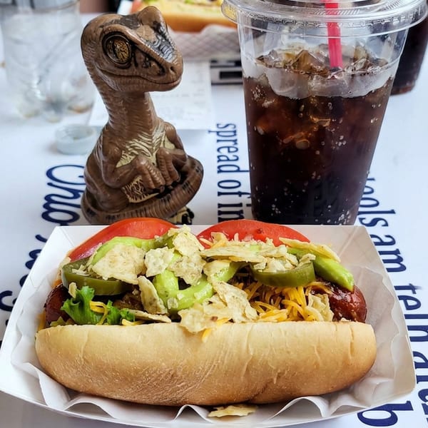 Loaded hot dog topped with avocado, tomato, shredded cheese, and crispy chips beside an iced cola and dinosaur figurine.