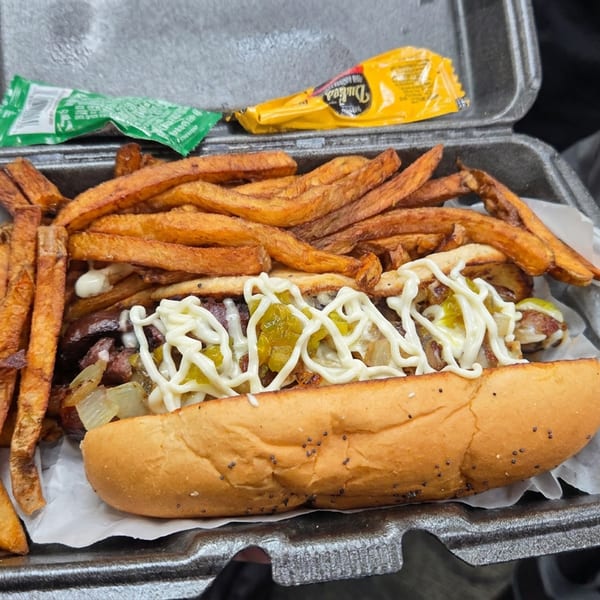 Steak hoagie topped with onions, peppers, and mayonnaise served with sweet potato fries in a takeout container.