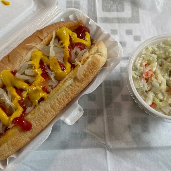 Hot dog topped with mustard, ketchup, and onions beside a container of creamy coleslaw in a takeout tray.