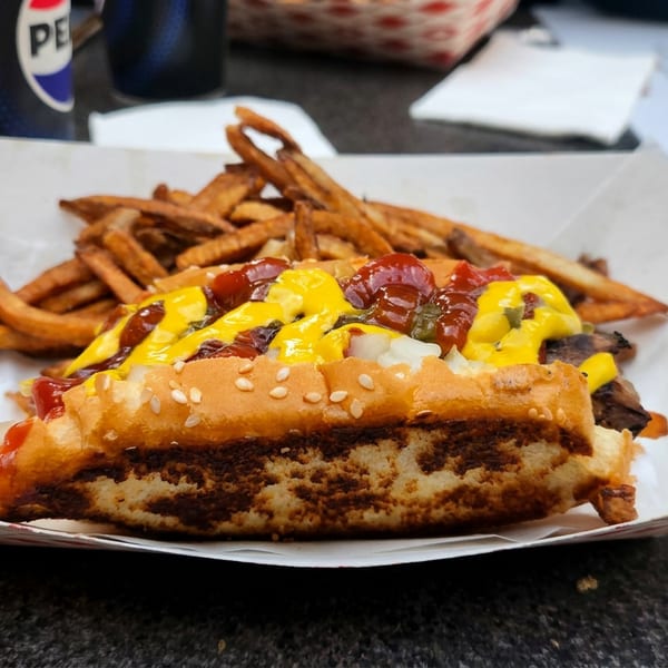 Cheeseburger topped with ketchup, mustard, onions, and pickles, served with fries on a paper-lined plate beside Pepsi drinks.