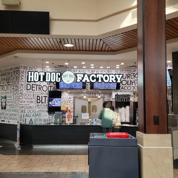 Hot Dog Factory counter in a food court, featuring bold city-themed wall graphics, menu screens, tables, and a customer ordering.